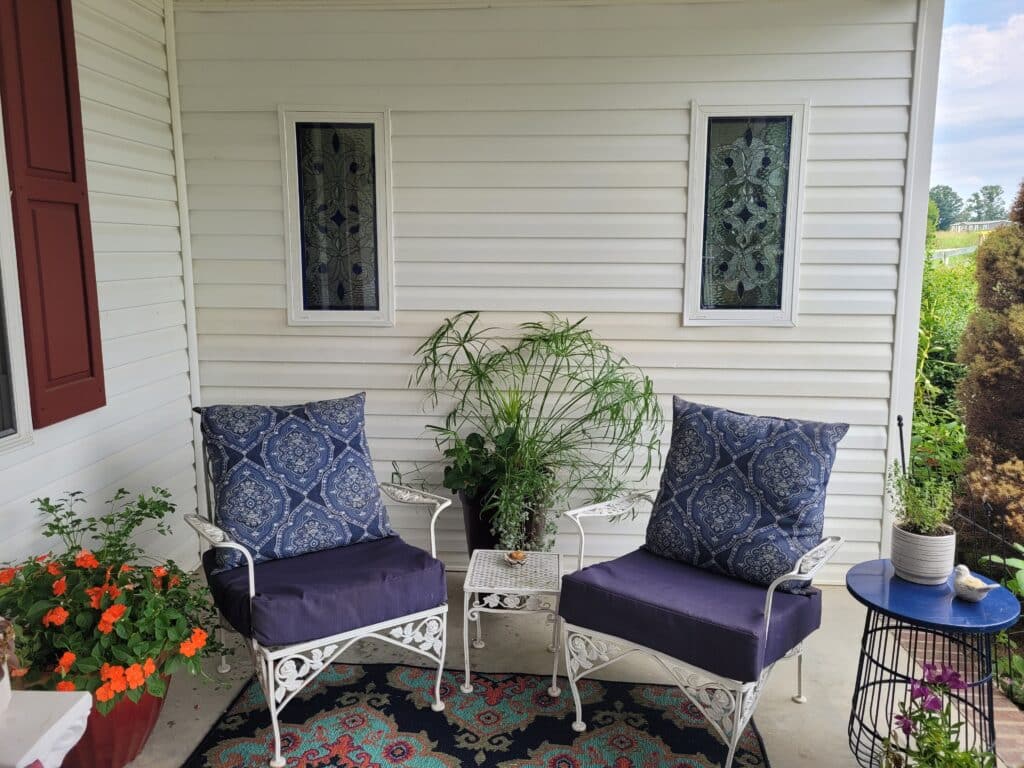 Two rectangular stained glass window inserts, each displaying an ornate, intricate scrollwork design in clear textured and beveled glass with dark blue (indigo) accent pieces, framed in white, and installed into a white-sided porch wall. The panels are positioned above two white patio chairs with blue cushions and patterned pillows, enhancing privacy and decor.