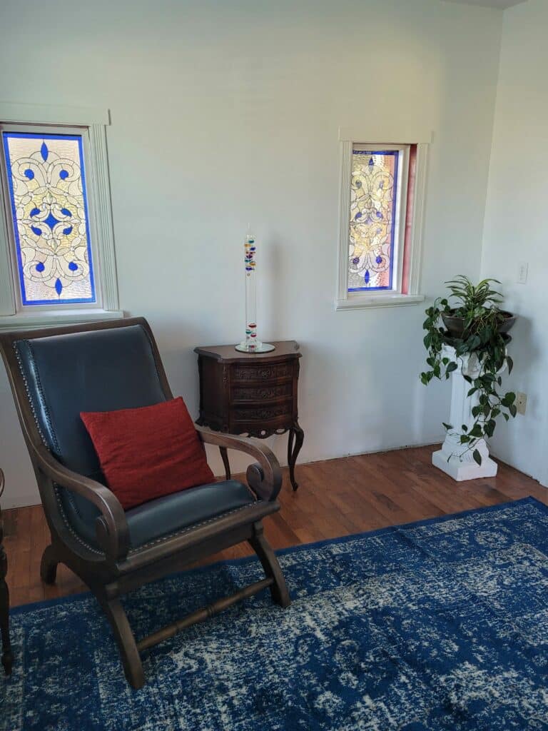Two rectangular leaded stained glass window panels showcasing an ornate traditional design. The design features rich royal blue fleur-de-lis motifs and flowing scrollwork made from clear, textured glass, set within white window frames in a room with white walls, hardwood floors, and a blue patterned rug. A dark wood rocking chair with a red cushion and an antique wooden side table are visible below the windows.