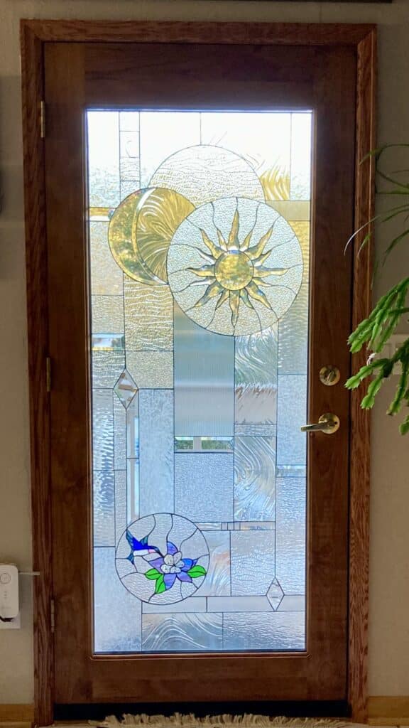 A full-length leaded stained glass door panel set in a rich wooden door. The panel features a complex design with various clear, textured, and beveled glass pieces. At the top, a prominent golden sunburst medallion and a textured golden crescent moon are visible. At the bottom, a circular medallion showcases a vibrant blue hummingbird feeding from a purple and blue flower with green leaves. The background consists of a mosaic of clear glass textures, including reeded, ripple, and geometric beveled diamond sections, providing privacy and light refraction.