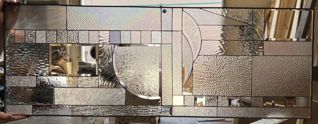 A rectangular stained glass panel featuring a complex, modern abstract geometric design. The panel is crafted from a diverse array of clear textured and beveled glass pieces, including ripple, stipple, hammered, reeded, and iridescent glass, with a prominent semi-circular element at its center. Light reflects dynamically off the varied surfaces, creating a shimmering effect.