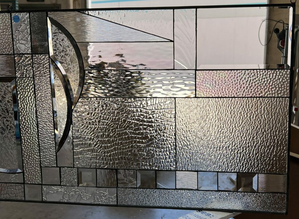 A rectangular, leaded stained glass panel with a modern abstract geometric design, crafted from various clear textured and beveled glass types. The panel is held vertically in a workshop, with a large garage door visible in the background.