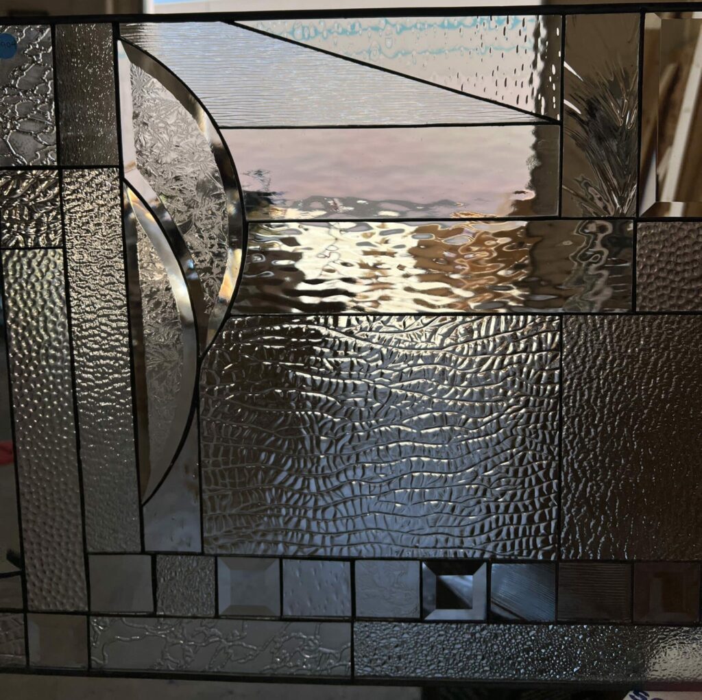 A rectangular, leaded clear stained glass panel featuring a contemporary abstract geometric design. It showcases a rich variety of clear and textured glass, including reeded, hammered, crocodile, and stippled patterns, along with prominent clear beveled glass accents and elegant curved lead lines.