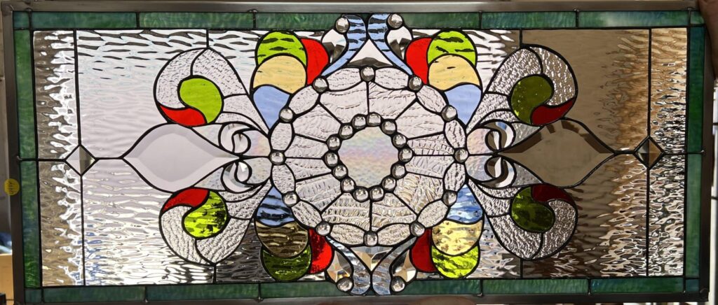 A rectangular leaded stained glass panel held by hands, featuring a central clear textured glass mandala-like medallion bordered by small round jewels. Symmetrically radiating from the center are organic and abstract shapes crafted from clear textured glass, accented with vibrant sections of red, lime green, light blue, and yellow glass. The entire composition is encased within a border of clear textured glass and a prominent green stained glass frame.