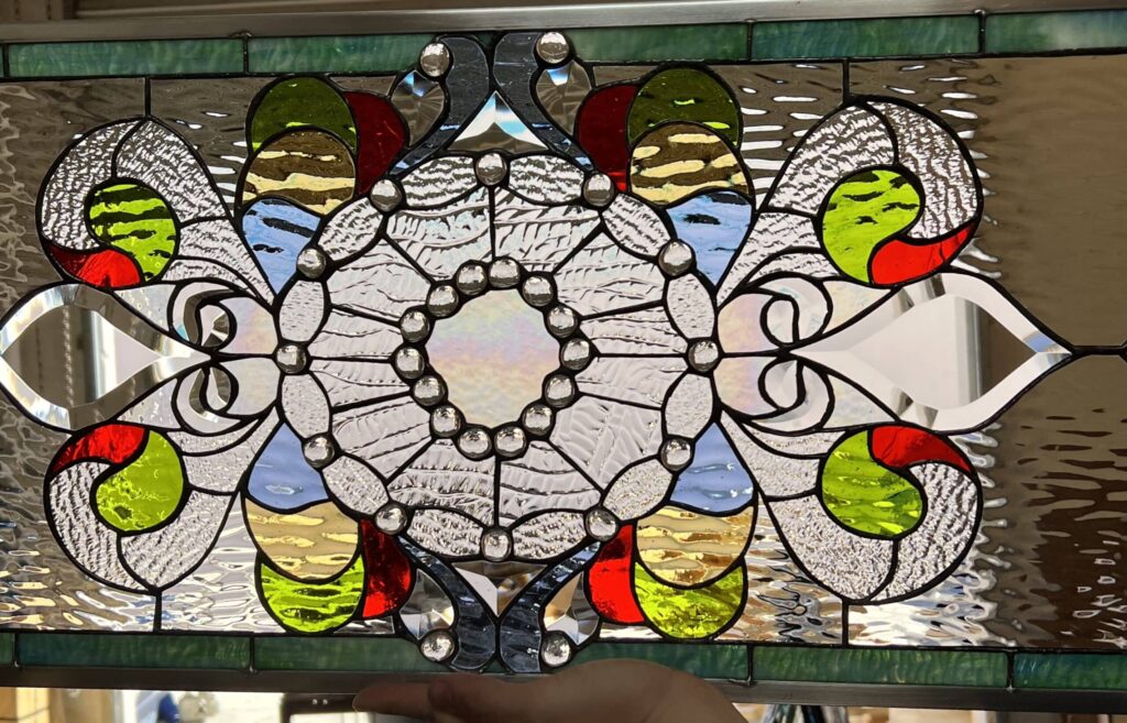 Rectangular leaded stained glass panel with a symmetrical ornate medallion design, featuring an iridescent clear center, clear beveled jewels, and various clear textured glass, accented with bright red, lime green, light blue, and amber colored glass. The panel is bordered by variegated green glass.
