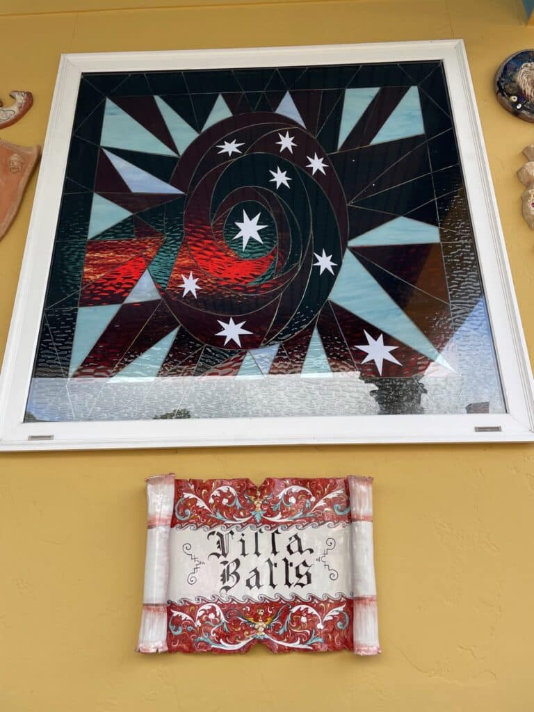 A rectangular stained glass panel in a white frame, featuring a dynamic spiral of dark and textured reddish-brown glass, dotted with white five-pointed stars. Light blue and clear textured glass triangles radiate outwards from the center, creating a cosmic or galactic effect.