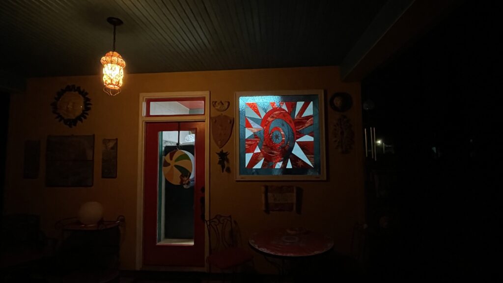 A brightly illuminated square leaded stained glass panel featuring a vibrant, abstract swirling design in reds, light blues, and whites. The design centers on a deep red spiral encircled by geometric shards and a small star, mounted on a warm yellow wall next to a red door.