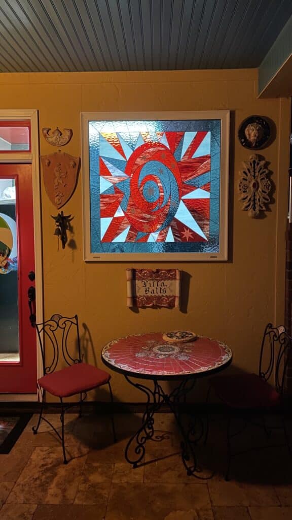 A rectangular stained glass panel featuring a vibrant abstract design with a central swirling motif in reds and oranges, surrounded by radiating triangular and trapezoidal pieces in light blue and clear textured glass. The panel, illuminated from behind, is set in a white frame on a golden-yellow wall above a small round table with a red mosaic top.