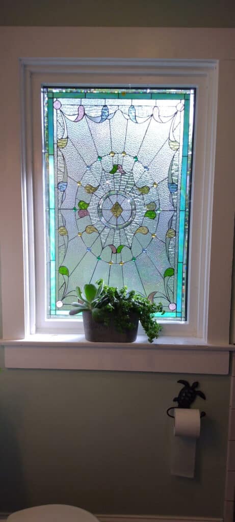 A rectangular stained glass window installed in a bathroom, featuring a central radiating pattern of clear textured glass (likely glue chip or hammered) with scattered accent pieces of green, blue, yellow, and soft pink colored glass. The design includes organic, leaf-like, and teardrop shapes, with a small central medallion. A potted succulent and trailing plant sit on the white windowsill below.