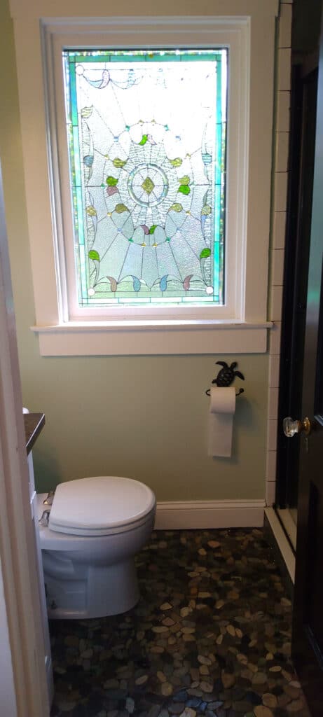 A rectangular stained glass window installed above a white toilet in a bathroom with light green walls. The leaded glass panel features a central circular medallion with a clear diamond, surrounded by swirling organic patterns made mostly of clear, textured privacy glass, accented with small elements in light green, blue, and soft pink/purple. The floor is covered in small, dark pebble stones, and a black turtle-shaped toilet paper holder is visible on the wall.