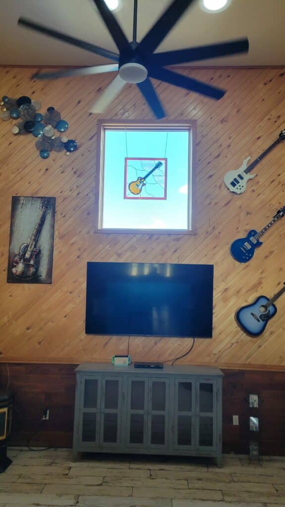 A rectangular stained glass window panel featuring a yellow electric guitar with a black neck, outlined in black leading. The guitar design is centered against a clear blue background, representing the sky, and enclosed within a red geometric border. The abstract leading lines around the guitar give it a modern, artistic feel.