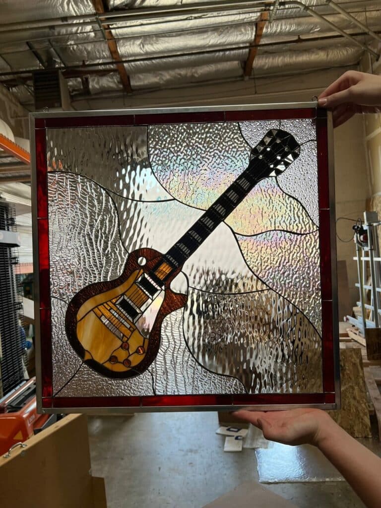 A square leaded stained glass panel depicting an electric guitar with a brown and amber body, black neck, and chrome details. The background is composed of various clear, textured, and iridescent stained glass pieces, all framed by a deep red border.