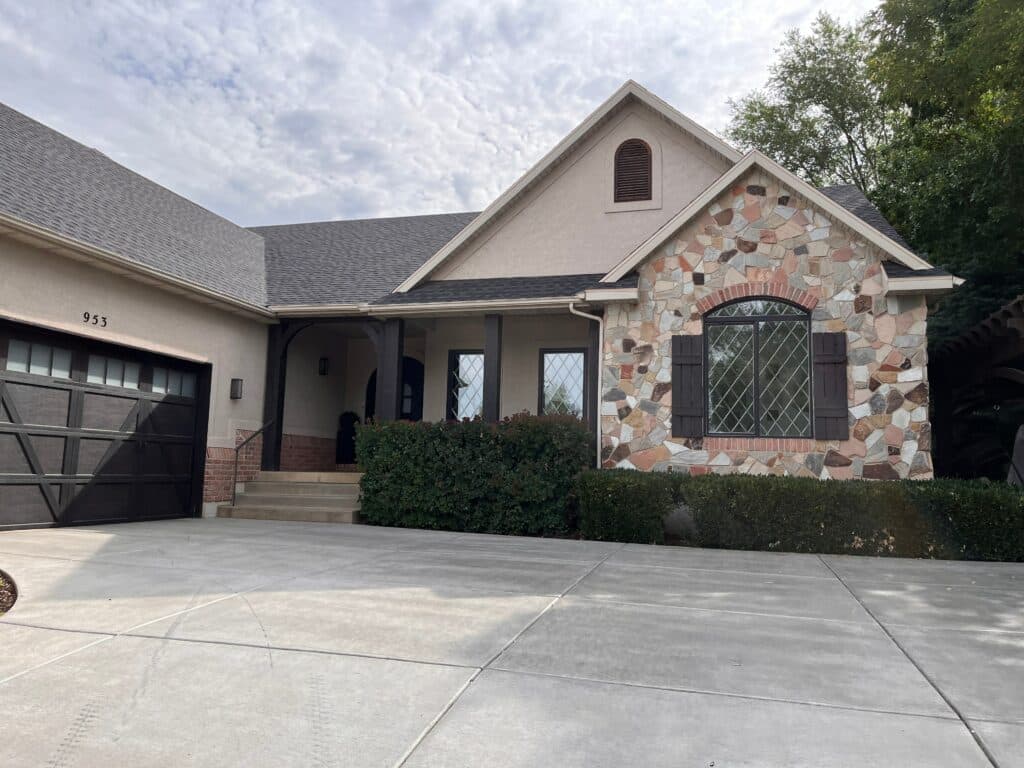 Exterior view of a residential home featuring stucco and natural stone cladding. The house has several windows, including a prominent arched window and two rectangular windows, all fitted with dark-framed leaded glass panels arranged in a classic diamond grid pattern. Dark wooden shutters adorn the arched window, and matching dark wood columns support a covered front porch.