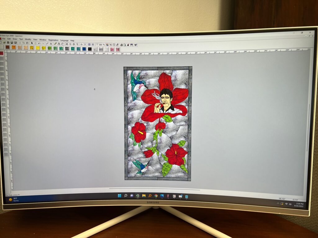 Digital stained glass design of Tony Montana from Scarface, smoking a cigarette, centered in a large red hibiscus flower, with two smaller red hibiscus blooms and two hummingbirds, all on a textured clear glass background within a leaded frame, displayed on a computer screen in Glass Eye 2000 software.