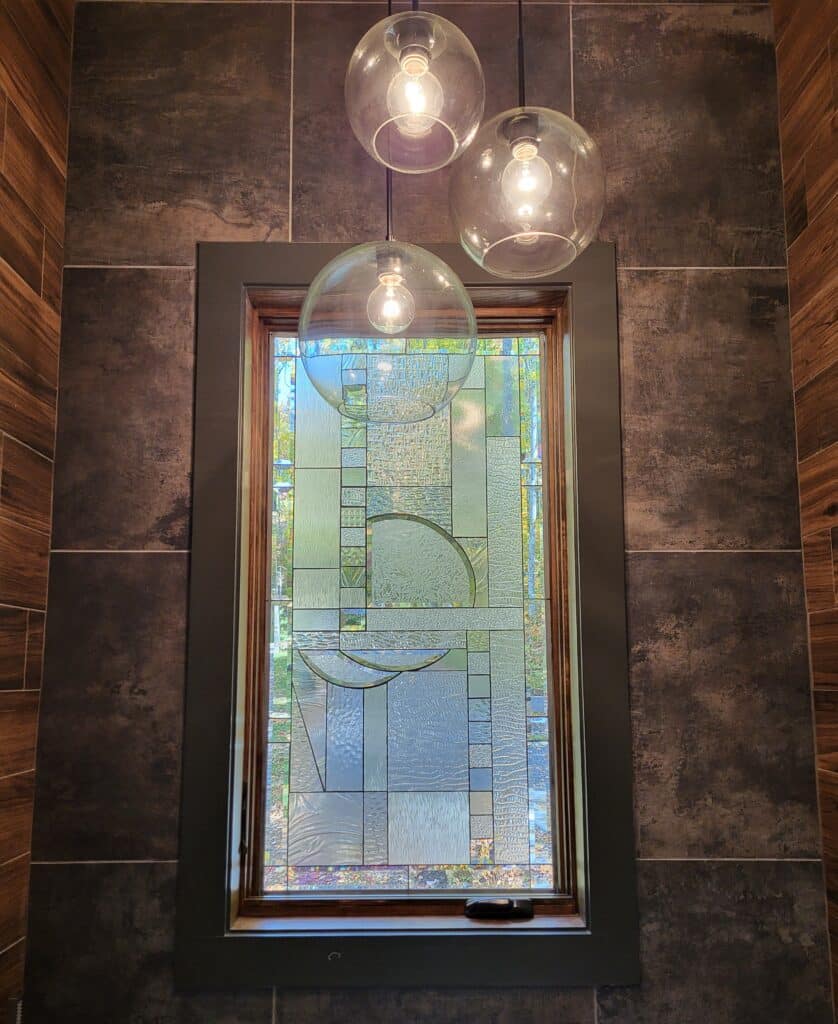 A rectangular stained glass window featuring an abstract geometric design made from clear, textured, and subtly iridescent glass, set within a dark wood frame. The window is flanked by dark tiled walls and illuminated by three spherical pendant lights.