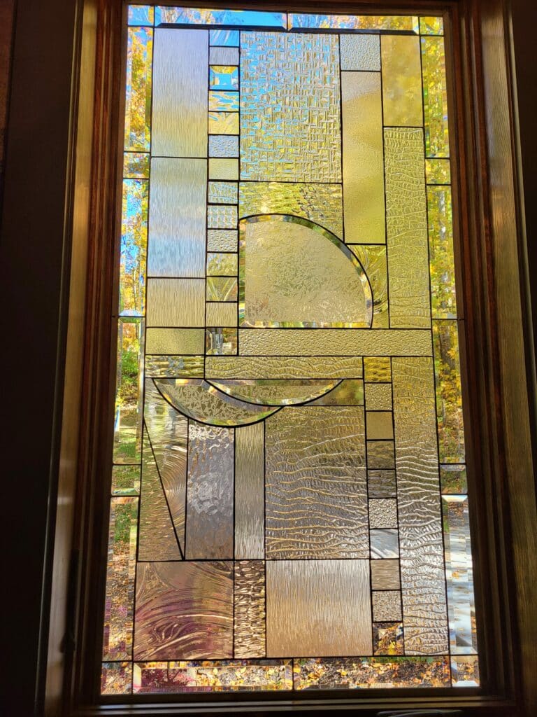 A rectangular, vertically oriented leaded stained glass window panel, showcasing an abstract geometric design. The panel features a rich variety of clear and lightly tinted textured glass, including wavy, reeded, hammered, and granular patterns, interspersed with clear beveled accents. Notable design elements include a large curved quarter-circle and two crescent shapes. The panel is framed in wood, with the warm colors of autumn foliage visible through the glass, casting golden and amber hues.