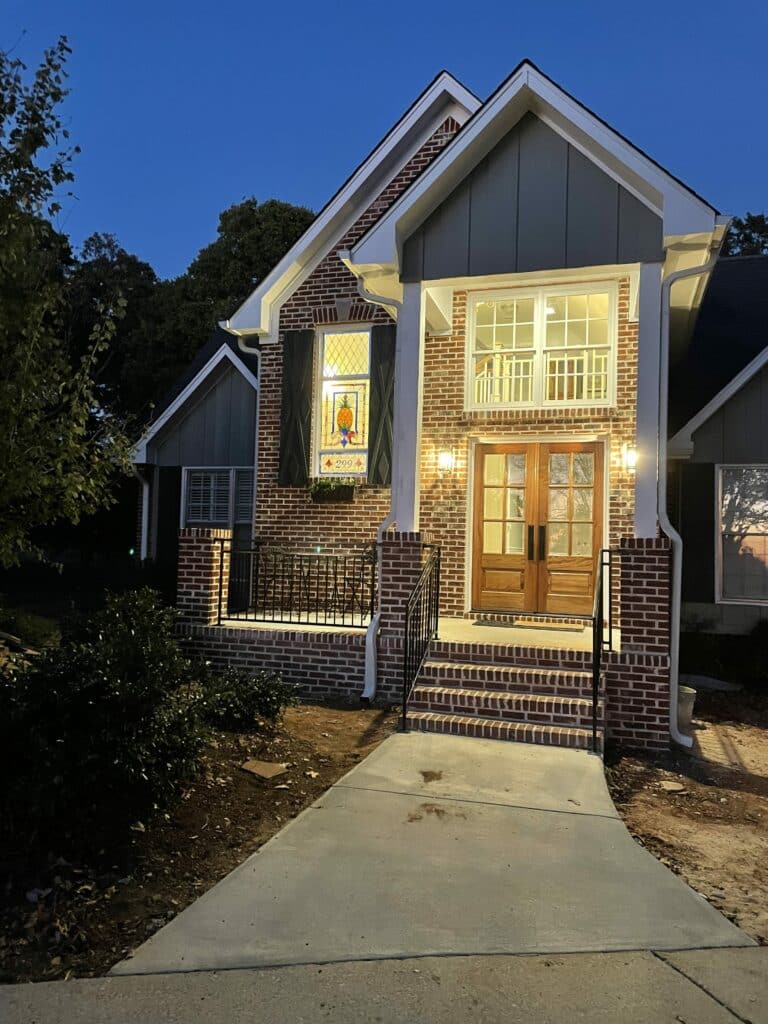 A warm, inviting brick home at dusk featuring a custom rectangular stained glass panel in a front window. The panel depicts a vibrant yellow and green pineapple, blue scrollwork, and the illuminated house number '299', flanked by dark decorative shutters. The porch has warm lighting, white columns, and wooden double doors.