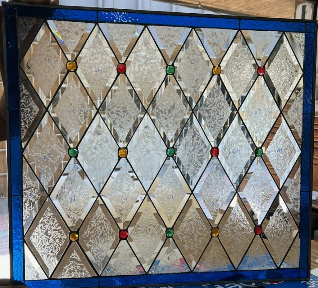 A rectangular leaded stained glass panel featuring a vibrant sapphire blue border. The main body consists of a diamond grid pattern of clear glass, alternating between reflective beveled diamonds and textured clear glass. Small, round red, yellow, and green jewel-like cabochons are interspersed at the diamond intersections.