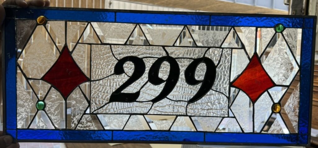 Rectangular leaded stained glass panel displaying the house number '299' in elegant black script, set against a background of clear textured and beveled glass. It features a vibrant cobalt blue border, two red diamond accents, and four small glass jewels (two amber, two emerald green) at design intersections.