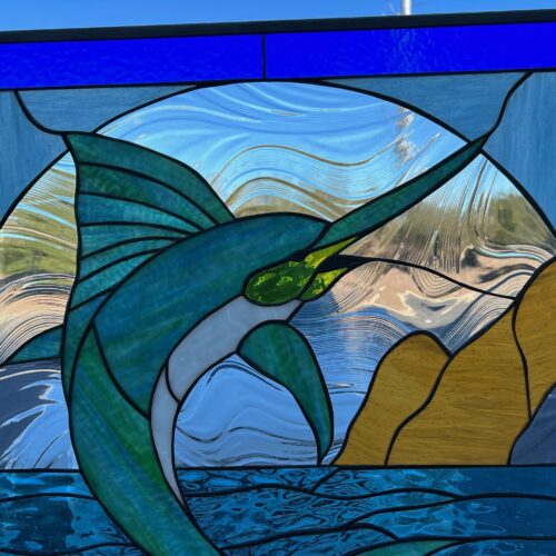 A rectangular stained glass panel depicting a vibrant blue-green marlin with a white belly leaping from textured blue ocean water, set against a background of golden-yellow landmasses and a swirling clear sky, framed by deep royal blue glass.