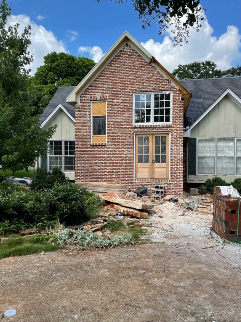 A rectangular stained glass panel featuring a detailed black leaded pineapple design, installed in a plywood-boarded window opening of a red brick house under renovation. Unfinished wooden double doors and construction debris are visible in the foreground.