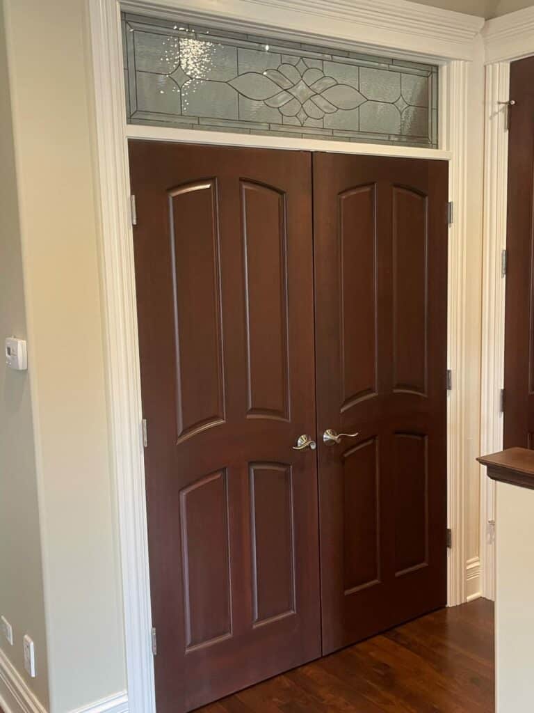 A rectangular leaded stained glass transom window, featuring a central clear beveled floral-geometric pattern surrounded by clear textured glass, installed above dark brown wooden double doors with crisp white trim.