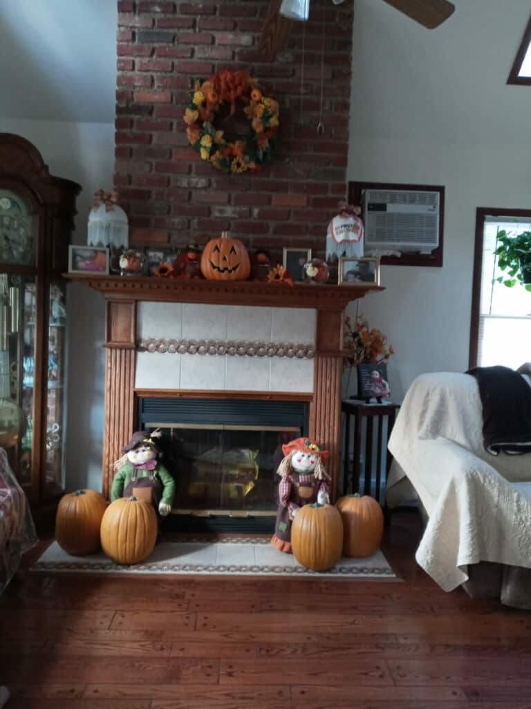 A brick fireplace with a wooden mantel, adorned with autumn decorations like a carved jack-o'-lantern, a fall wreath, and two scarecrow dolls with pumpkins. A grandfather clock is on the left, an armchair on the right, and hardwood floors below.