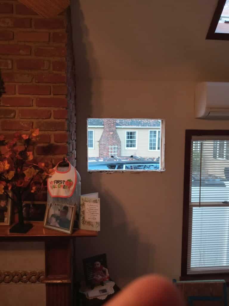 A rectangular opening cut into a light brown interior wall, revealing a portion of a neighboring house with beige siding and a prominent brick chimney. To the left, a section of an interior brick column is visible, along with a shelf holding a 'My First Halloween' bib, framed photos, and artificial autumn leaf decor. A window with horizontal blinds is on the right, and the bottom right corner of the image is blurred by a finger.