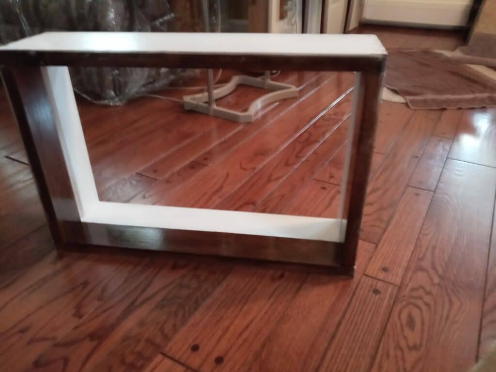 A rectangular, empty frame featuring a dark brown wooden exterior and a contrasting white interior border, designed for a custom glass insert, resting on a polished hardwood floor.