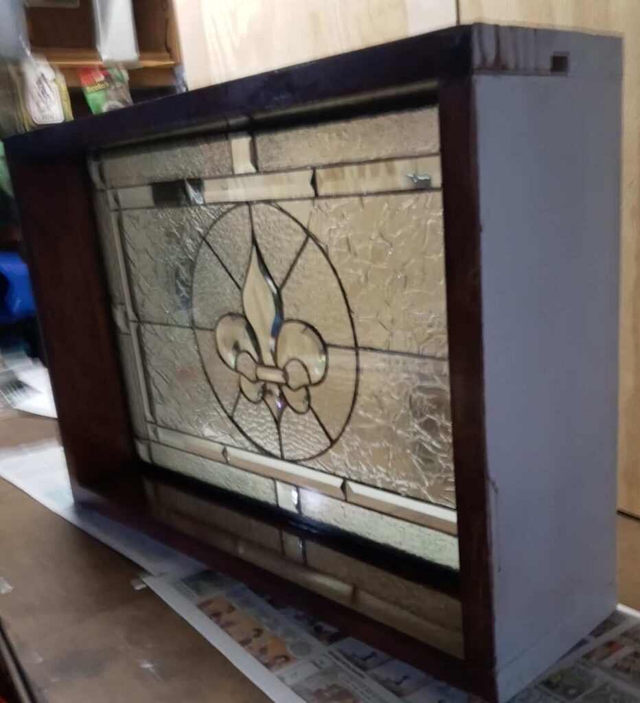 A rectangular leaded stained glass panel displaying a central clear beveled fleur-de-lis, encircled by clear textured glass and clear rectangular bevels, set within a dark wooden frame.