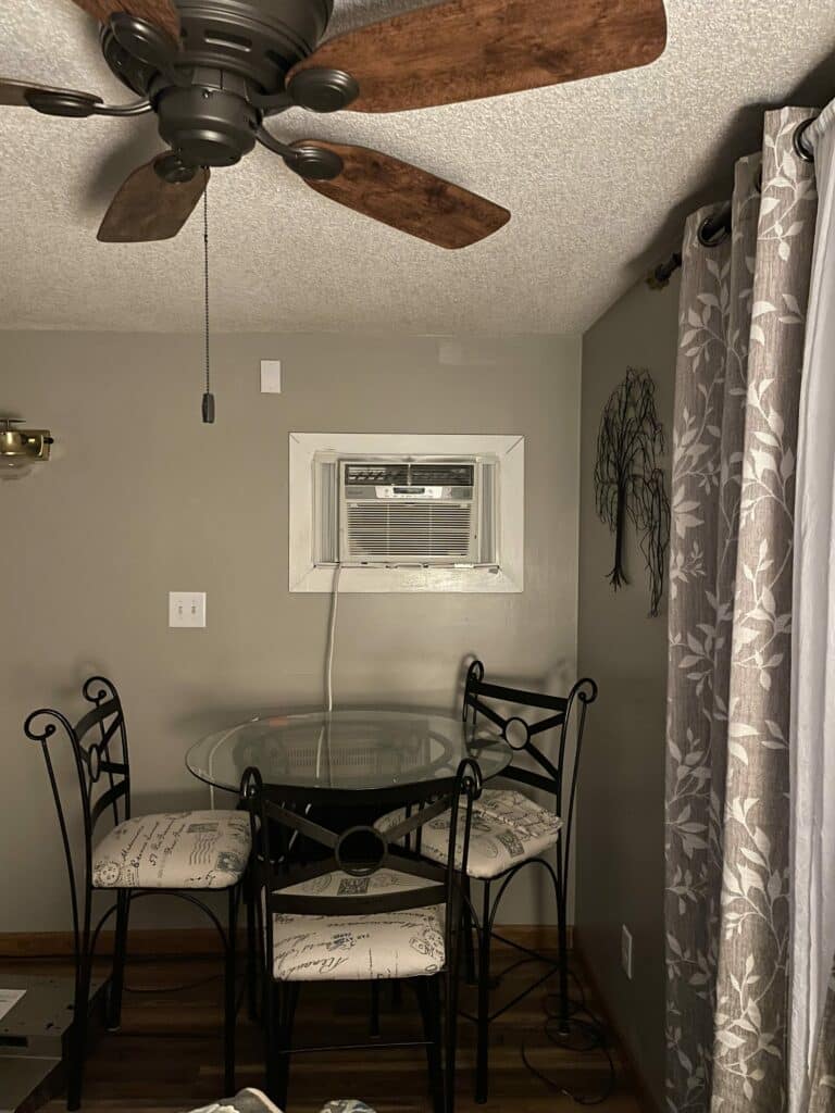 A rectangular clear beveled stained glass panel, designed to provide privacy and light while fitting into a standard window opening. The panel features a geometric leaded design with various clear textured and prismatic beveled glass pieces. It is depicted as an ideal replacement or enhancement for a window above a dining table in a kitchen nook setting.
