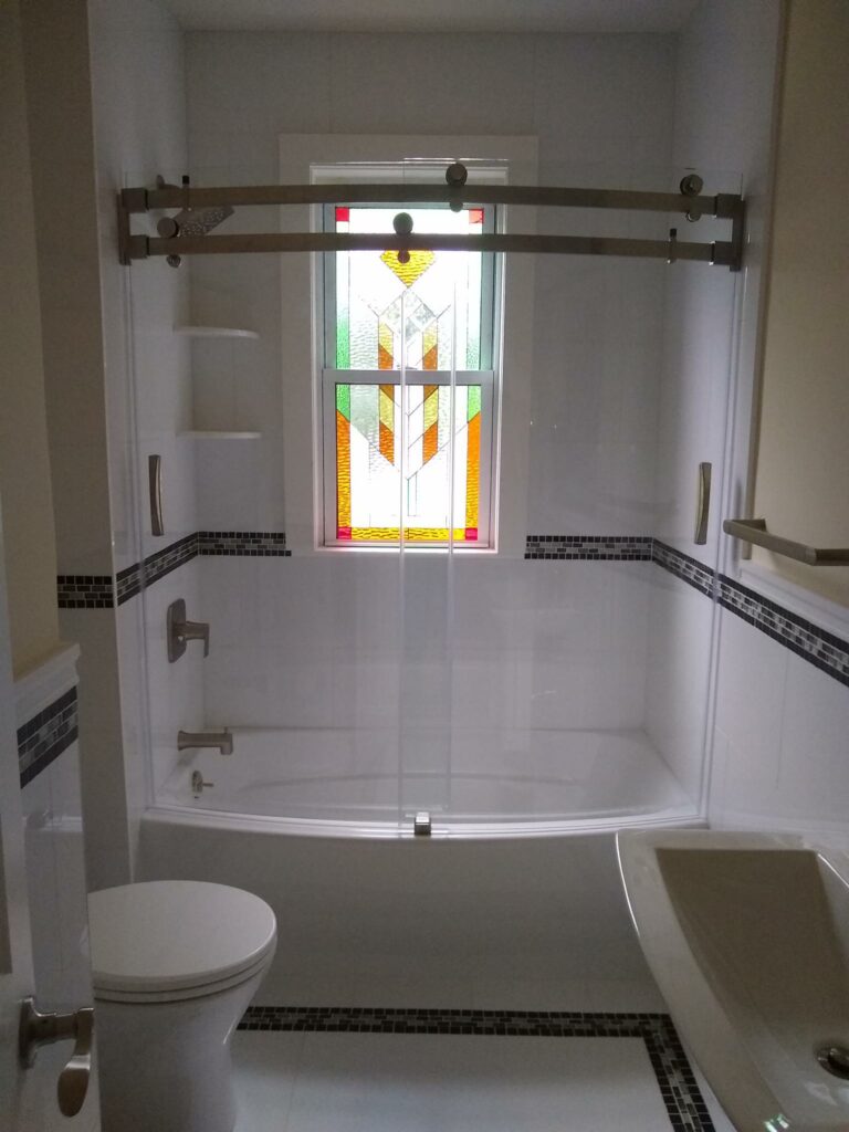 A rectangular stained glass window installed above a white bathtub in a modern bathroom. The leaded glass panel features a vibrant, abstract geometric design with clear, green, orange, and red glass, providing privacy.