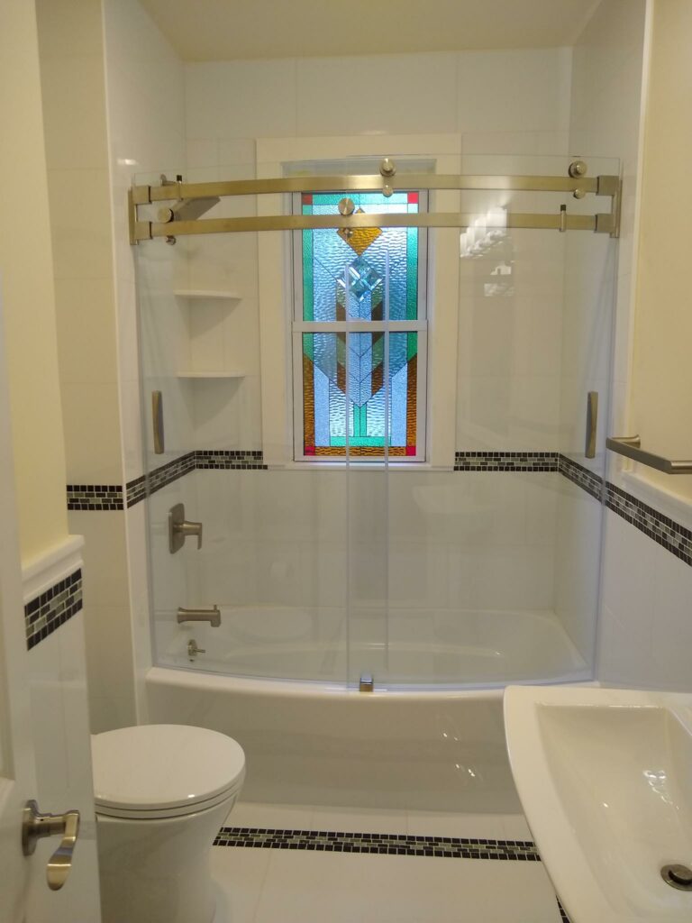 A rectangular stained glass window with a contemporary geometric pattern featuring blues, greens, and amber textured glass, installed above a white bathtub in a modern bathroom.