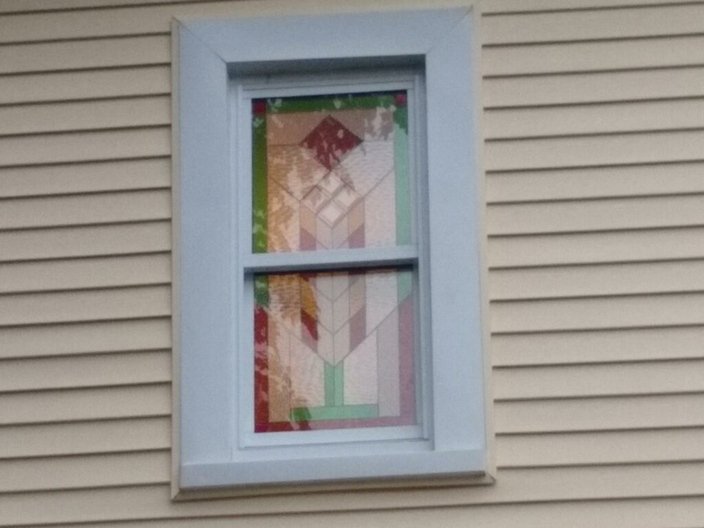 A rectangular leaded stained glass window panel, showcasing an abstract geometric Prairie-style design with segments of clear, textured, light green, amber, and muted red glass, installed in a light blue window frame on a house with beige siding.