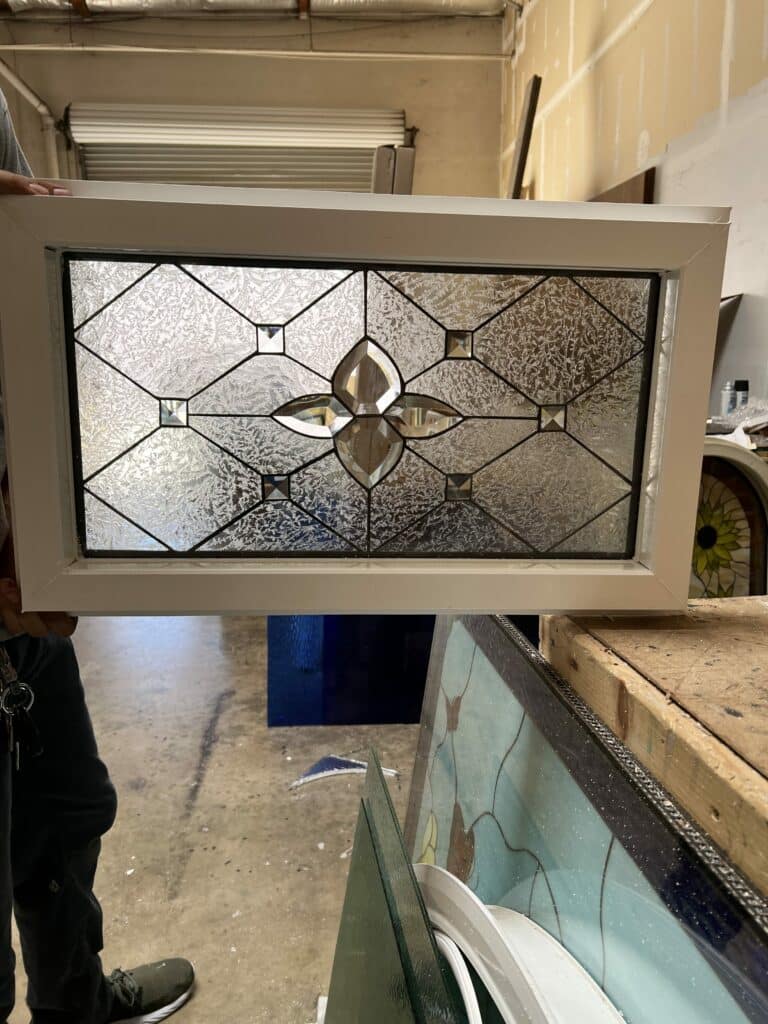 A rectangular leaded stained glass panel set in a white frame, showcasing a central clear beveled four-petal motif, flanked by textured clear glass with a delicate fern-like pattern, and accented with smaller square beveled pieces.
