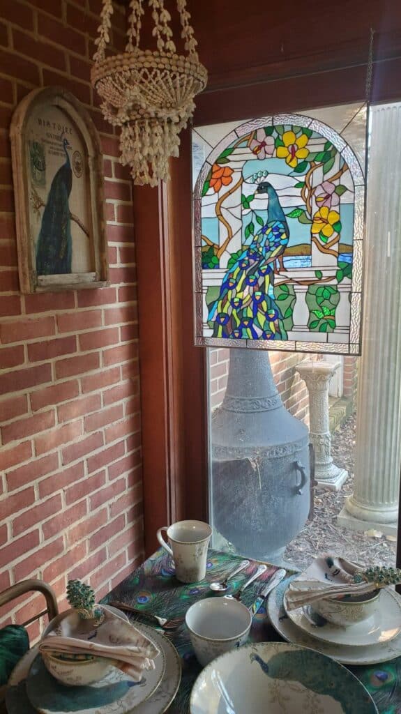 An arched stained glass panel depicting a colorful peacock with blue and green tail feathers, perched amidst pink and yellow flowers on branches, against a light blue sky. The panel features a decorative leaded design and a textured clear border.