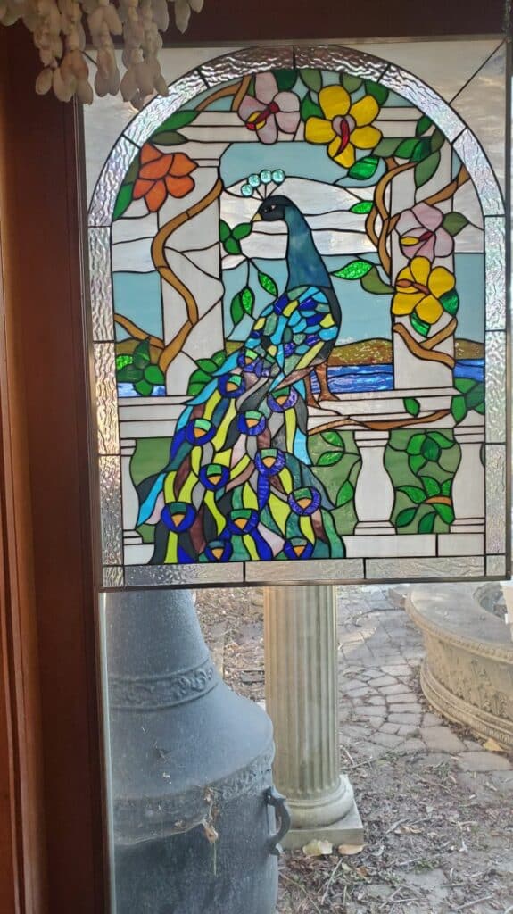 An arched, leaded stained glass panel depicting a vibrant blue and green peacock with a fanned tail, perched on a white balustrade amidst a lush garden with colorful pink, yellow, and orange flowers, green foliage, and a light blue background.