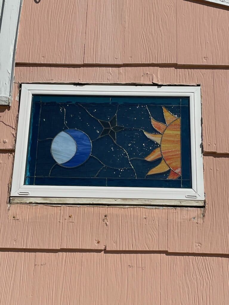 A rectangular stained glass window panel depicting a bright orange sun and a blue crescent moon against a dark blue, starry sky, installed in a pink shiplap siding wall.