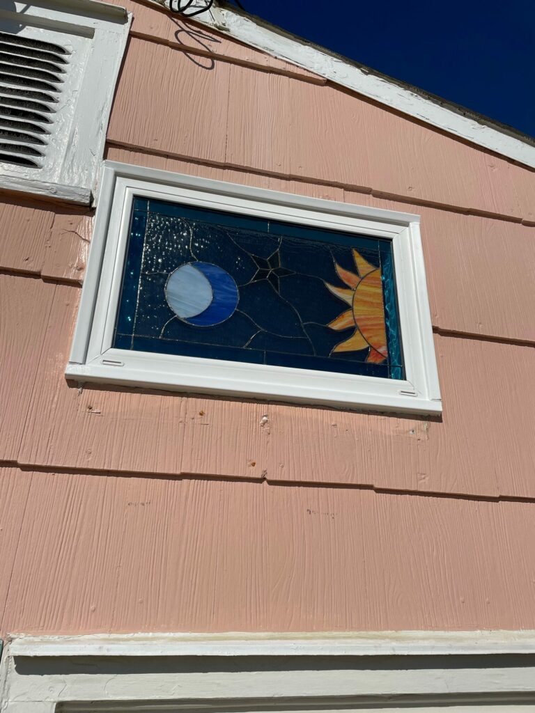 A rectangular stained glass window panel installed in a white frame on a textured pink house. The panel depicts a celestial scene with a crescent moon on the left and a stylized sun on the right, both set against a dark blue background with textured clear glass elements representing stars. The moon is crafted from light and dark blue swirled glass, and the sun features bright yellow, orange, and red-orange radiating rays.