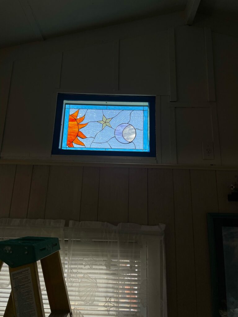 A rectangular stained glass panel depicting a celestial scene. On the left, a vibrant orange sun with pointed rays. In the upper center, a small yellow five-pointed star. On the right, a white and purple crescent moon. The background is composed of light blue clear textured glass, separated by dark lead lines, suggesting an ethereal sky. The panel is set into a wall opening, surrounded by dark trim.