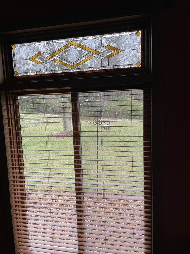 A rectangular leaded stained glass transom panel featuring an abstract geometric design with clear textured glass and amber diamond accents, installed above a window overlooking a snowy winter yard.