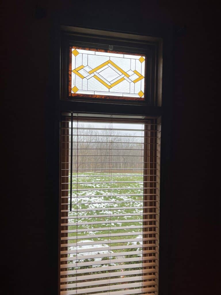 A rectangular leaded stained glass transom panel installed above a window with wooden blinds, showcasing a central geometric pattern of clear textured glass and golden yellow diamond shapes, surrounded by a border of rich amber textured glass.