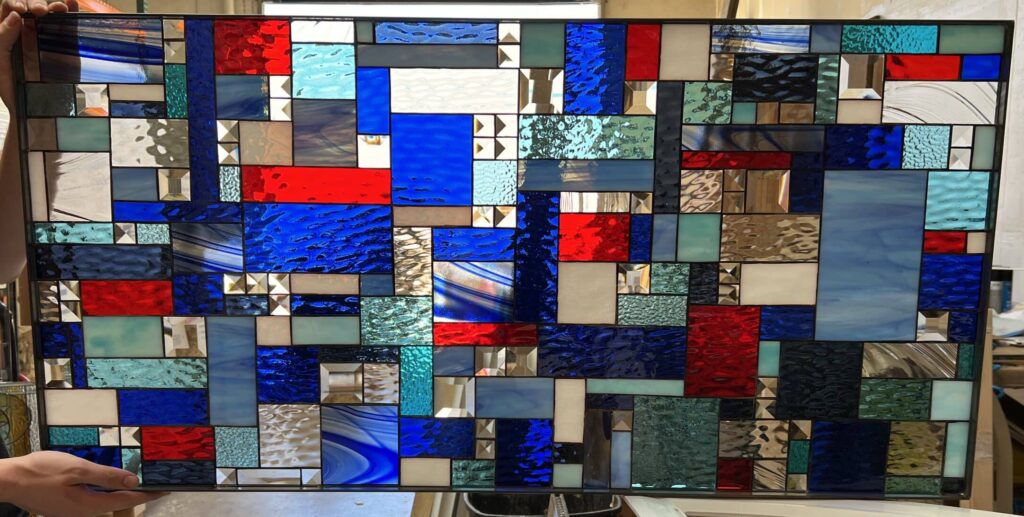 A rectangular, leaded stained glass panel displaying a vibrant abstract geometric design. It features a mosaic of blue, red, clear, and teal art glass pieces with various textures like ripple, hammered, and swirled, accented by numerous clear beveled squares and rectangles.
