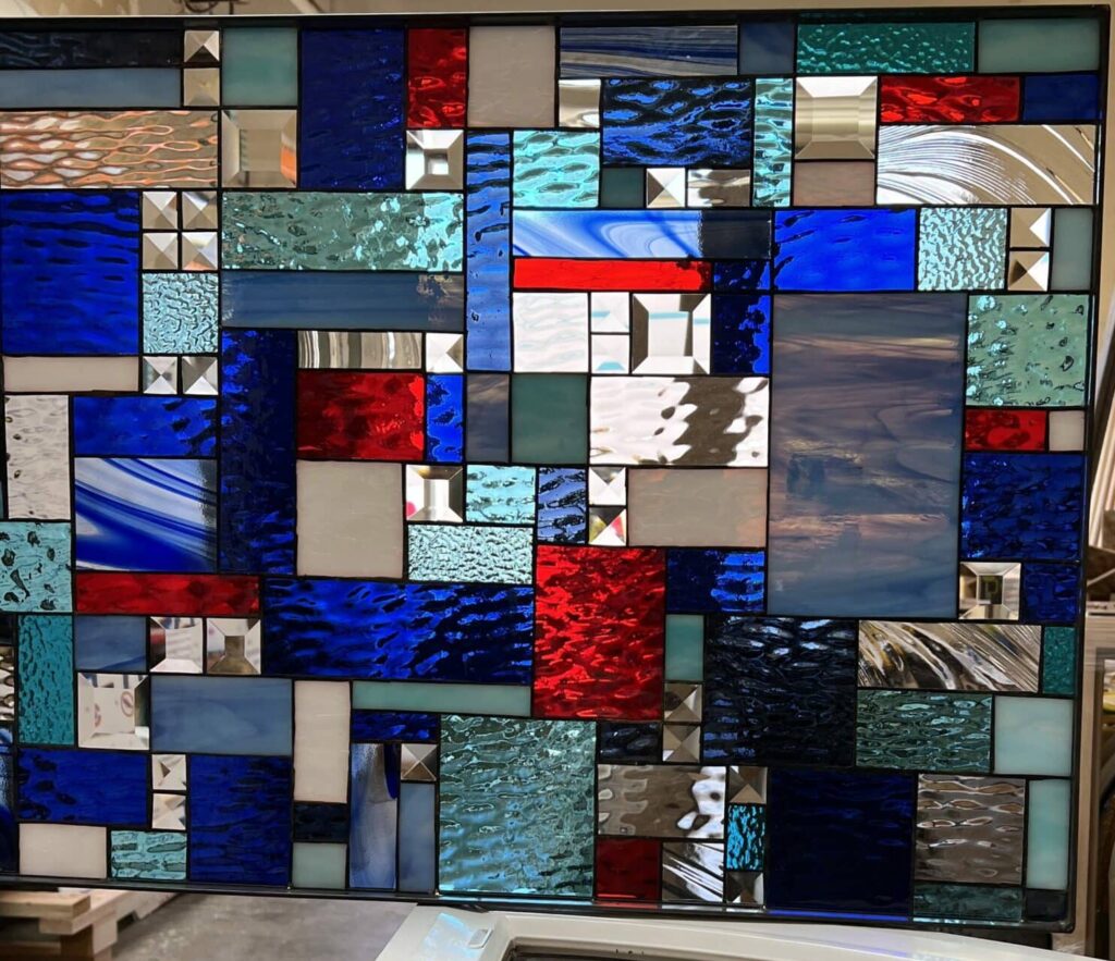 A large rectangular stained glass panel showcasing a vibrant abstract geometric design, featuring blocks of deep blue, bright red, clear textured, and white glass, interspersed with faceted clear bevels. The panel is set in a workshop, with a curved window in the foreground reflecting a separate stained glass panel depicting a yellow sunflower.
