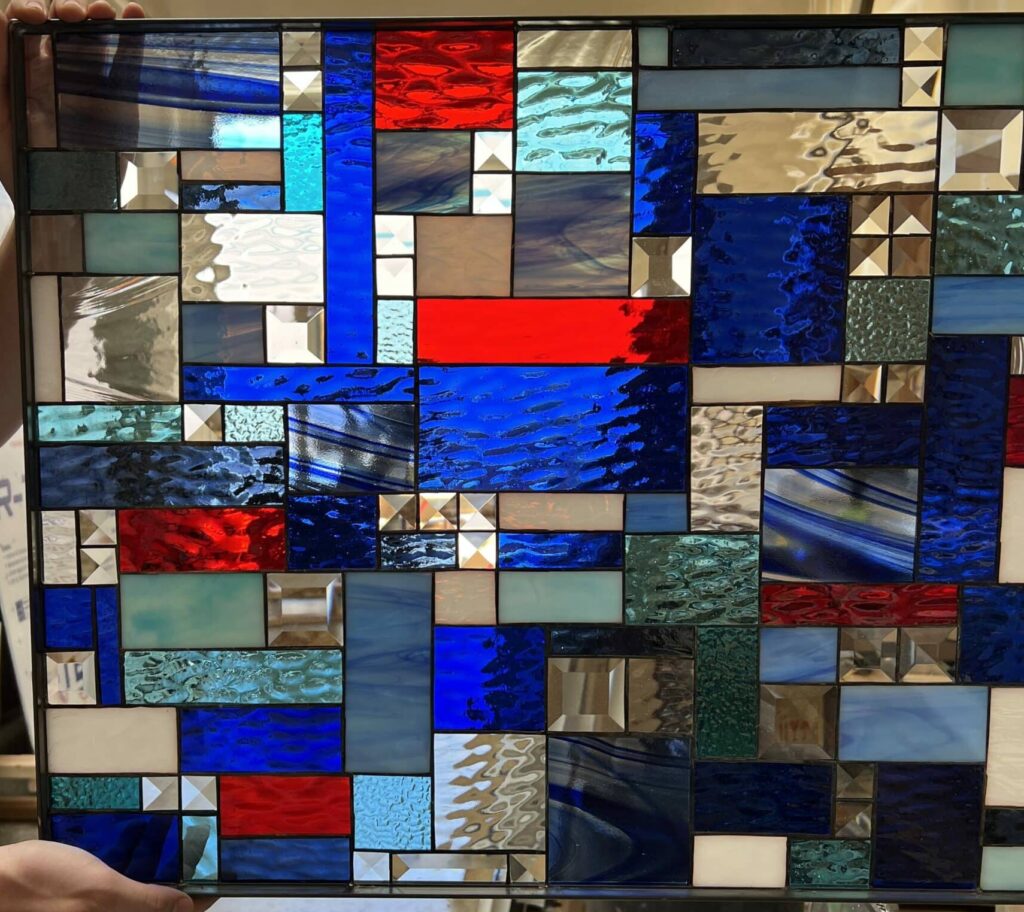 A rectangular leaded stained glass panel held by two hands, displaying a vibrant abstract geometric pattern. The design incorporates diverse textures of blue, red, clear, and white art glass, accented with multifaceted square and rectangular bevels.