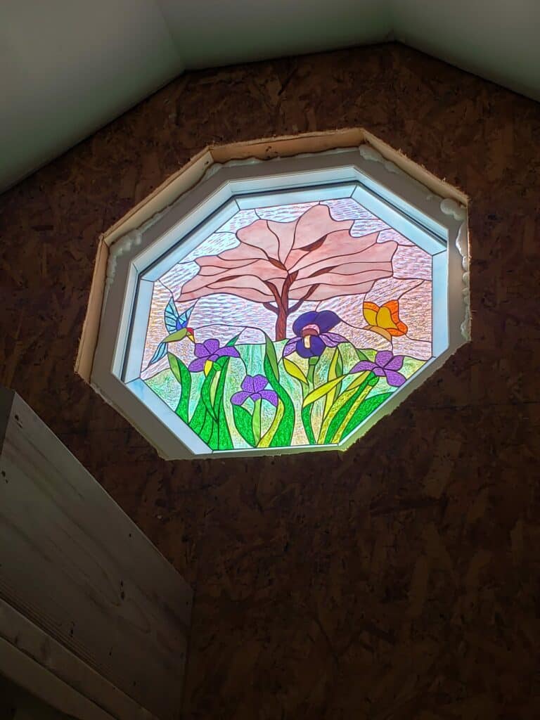 An octagonal, white-framed insulated window unit featuring a vibrant leaded stained glass panel. The design depicts a lush garden scene with a light pink-leaved tree in the background, rich green foliage and purple iris flowers in the foreground, a blue hummingbird hovering on the left, and a yellow butterfly on the right.
