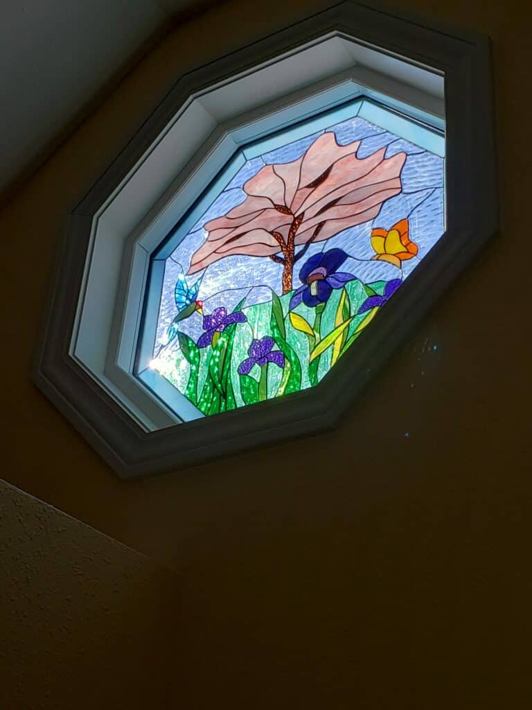 An octagonal leaded stained glass window, framed in white, depicting a vibrant garden scene. It features purple irises, green textured foliage, a large peach-colored tree with a brown trunk, a blue hummingbird in flight, and a yellow butterfly. The background is a light blue, textured sky.