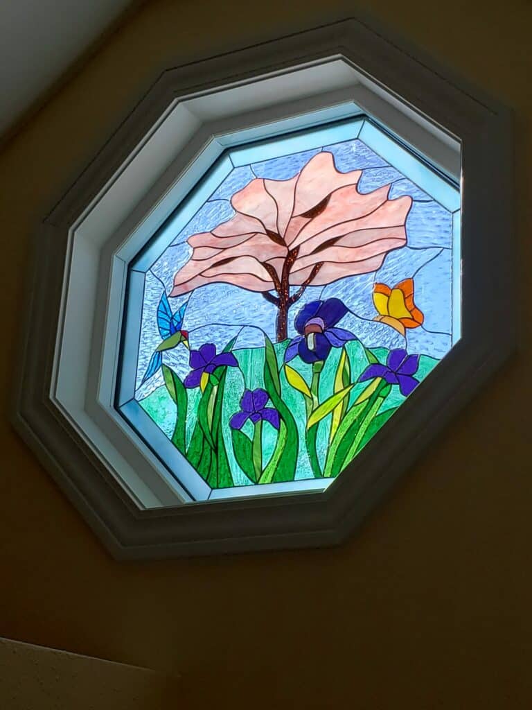 An octagonal stained glass window depicting a vibrant natural scene. It features a tree with pinkish-orange leaves, several purple iris flowers with green stems, a blue hummingbird, and a yellow butterfly, all set against a light blue textured sky and green ground, encased in a white frame.