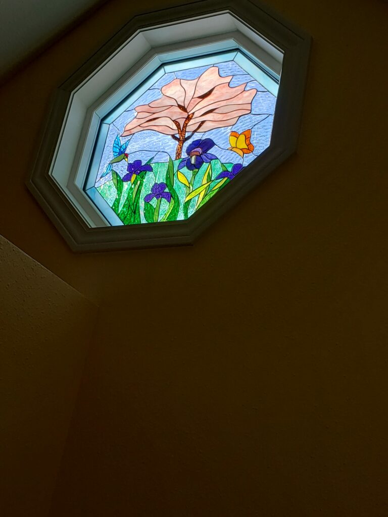 An octagonal, leaded stained glass window panel depicting a vibrant nature scene with a light-orange tree, purple irises, a blue hummingbird, and an orange butterfly against a textured light blue background, set in a white frame.