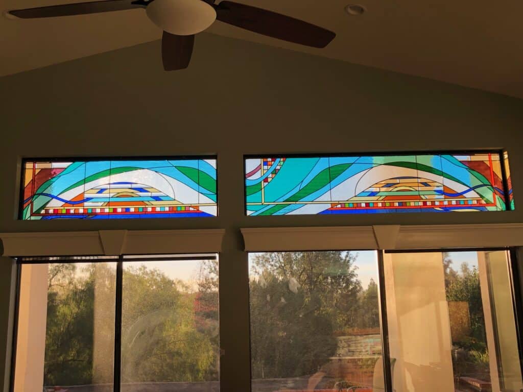 Two side-by-side rectangular stained glass transom windows, featuring an abstract, flowing design with large wave-like forms in blues, greens, and white, accented by smaller horizontal bands of colorful geometric squares in red, orange, yellow, and blue. The panels are installed high on a wall above clear windows looking out to trees.