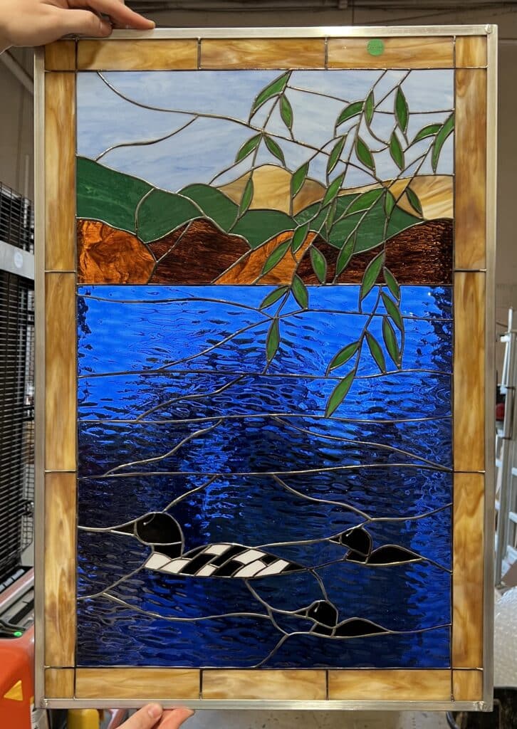 A rectangular, leaded stained glass panel depicting a serene lake scene with light blue sky, green and brown rolling hills, and deep textured royal blue water. Three black and white loons swim in the water, while green leafy branches hang from the top. The panel is bordered with variegated amber glass and framed in silver-toned metal.